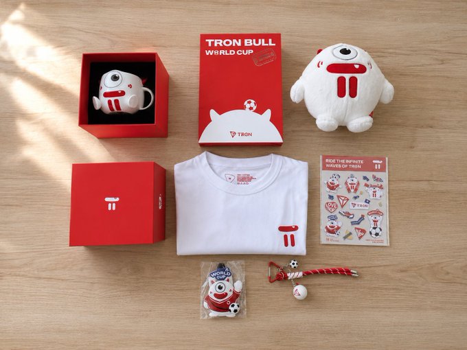 TRON-branded merchandise laid out on a wooden surface, including a red "TRON BULL WORLD CUP" box, a white plush mascot, a t-shirt, a mug, stickers, a small toy, and a decorative cord.