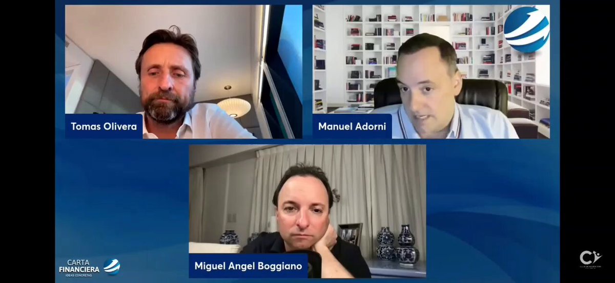 The image shows three men sitting at a table, each with their own laptop open in front of them during an online meeting or conference call. The men are facing the camera, appearing engaged in discussion or presentation. The background features a blue wall adorned with bookshelves filled with various books and other items, suggesting that they might be discussing business or work-related topics.