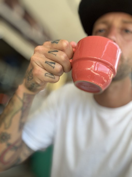 A tattooed man wearing a black cap and white shirt drinks from a bright coral-red ceramic mug, capturing a casual morning coffee moment that ties into the post about his habit of starting the day with 2-3 coffees.