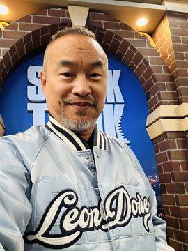 A man wearing a light blue "Leon Dore" varsity jacket takes a selfie in front of a brick archway with partially visible blue lettering behind him. The image accompanies a post about crypto trader "Machi" turning $152K into $12.72M in three days following nearly 500 liquidations.