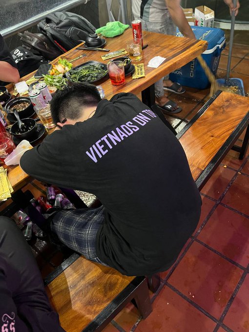 A person resting their head on a wooden table in what appears to be a restaurant, wearing a black shirt with 'VIETNADS ON TOP' printed in purple text.