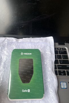 A green Trezor Safe 1 cryptocurrency hardware wallet resting on its packaging on a desk next to a laptop.
