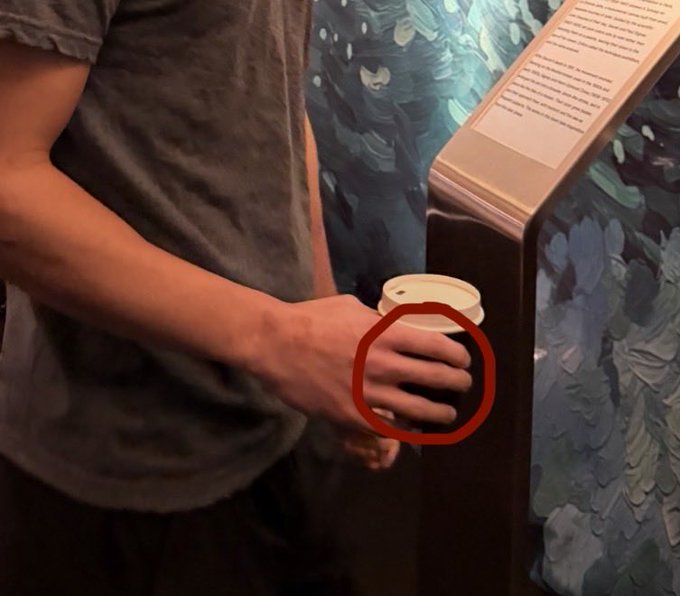 A person in a grey shirt holds a paper cup, with a red circle highlighting their unusually elongated or deformed fingers.
