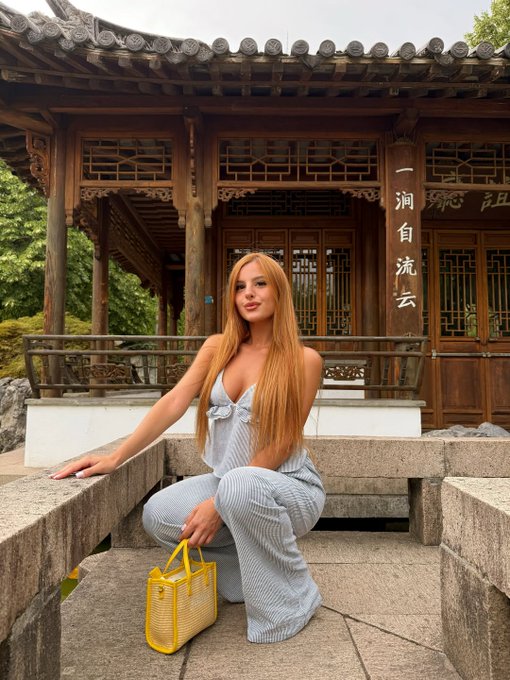 Woman with long red hair in a light blue ribbed outfit crouching on stone steps in front of a traditional Chinese pavilion with wooden lattice windows and Chinese calligraphy, holding a yellow handbag.