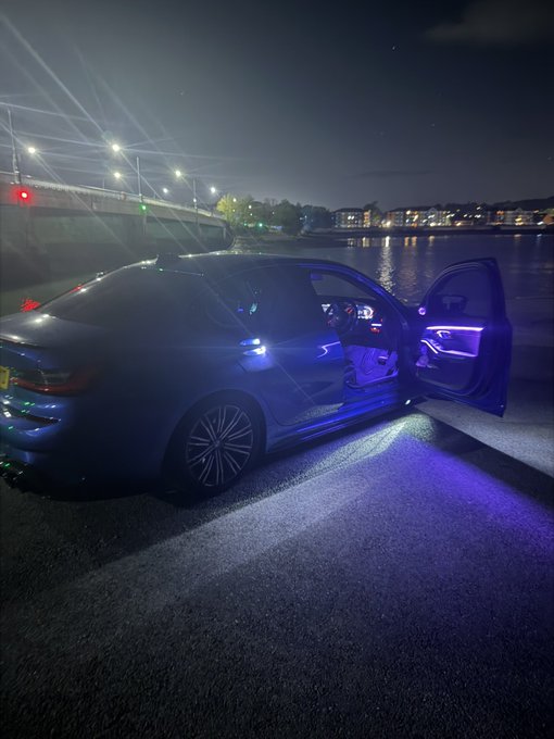 A BMW sedan with its doors open showcases vibrant purple ambient interior lighting, parked by a waterfront under a bridge lit by streetlamps at night, exemplifying the premium "GM bros" vibe of a luxury cruise setting.