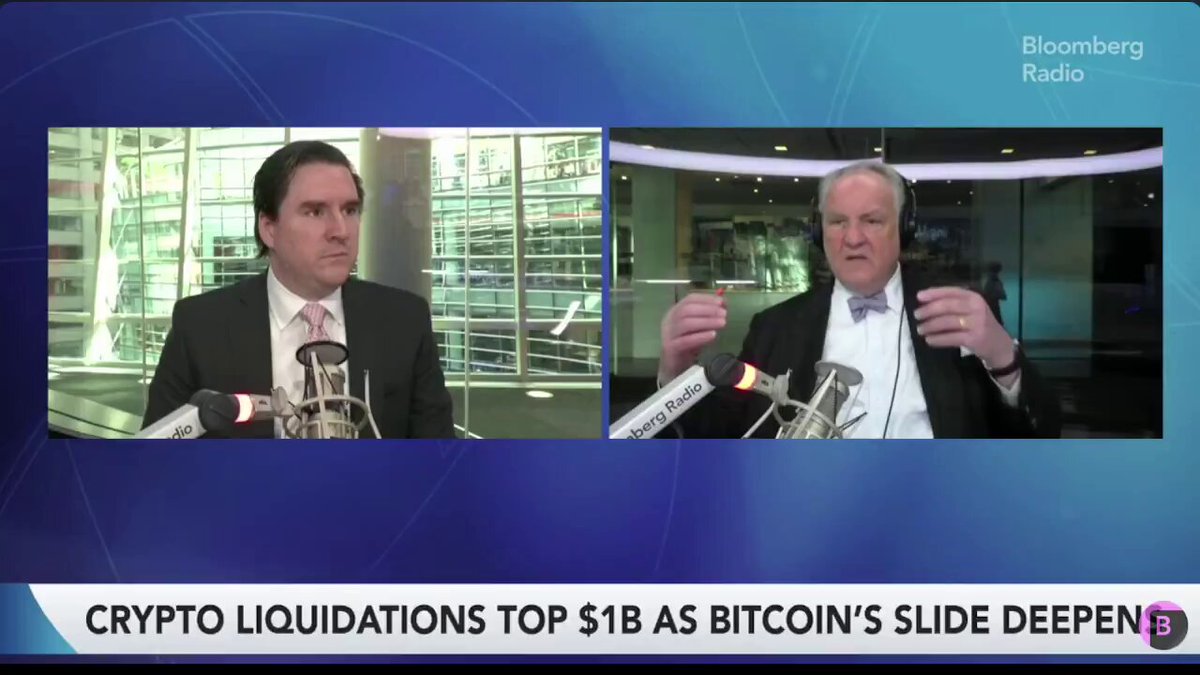 The image shows two men sitting at a desk, with one man wearing a suit and tie while the other has a microphone. They are both talking about Bitcoin and its value. The background of the image features a blue sky and buildings, suggesting that they might be in an office setting or a newsroom.
