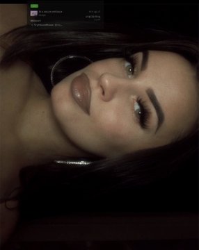 A selfie of a woman with dark hair, dark lipstick, and a nose ring in a low-light setting, appearing upside down with a small chat overlay in the upper left.