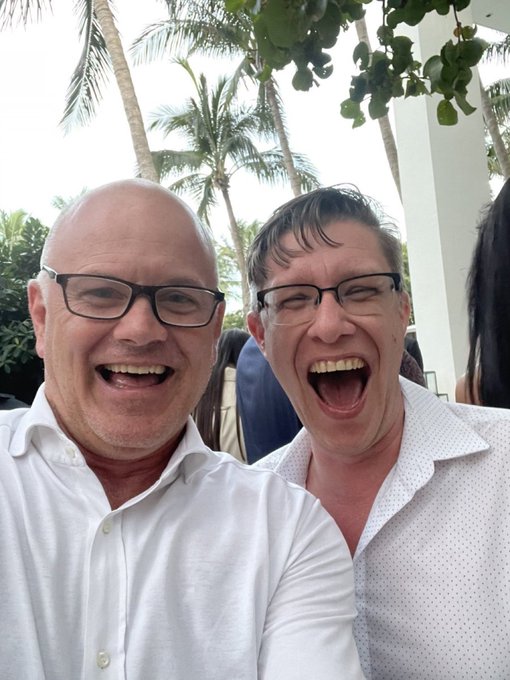 Two men wearing glasses laugh heartily while taking a selfie outdoors, with palm trees visible in the background.