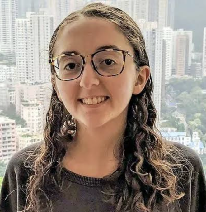 A young woman with glasses smiles at the camera, with a dense cityscape of high-rise buildings visible through the window behind her.

The image appears unrelated to the post's subject matter, which discusses the CFTC's resolution of enforcement actions against Caroline Ellison and Gary Wang, including five-year trading bans, as neither individual is visible in the photo.