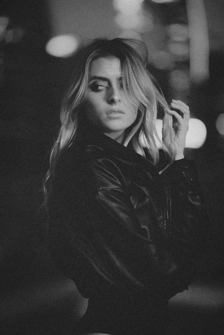 A black and white portrait of a young woman with wavy blonde hair wearing a dark jacket, looking toward the camera with her hand raised to her hair.