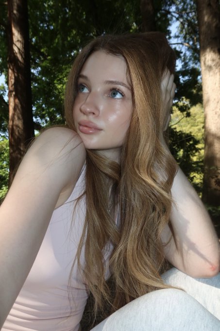 A portrait of a young woman with long brown hair looking upward, wearing a light pink sleeveless top while sitting outdoors in a forest setting.