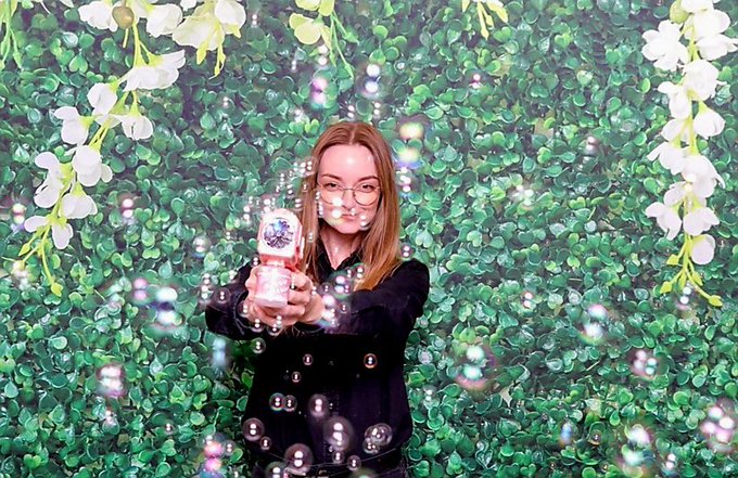 A young woman with glasses points a bubble gun toward the camera, surrounded by floating bubbles against a lush green foliage backdrop with white flowers framing the scene.