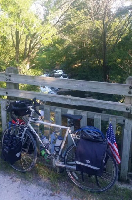 A touring bicycle equipped with rear panniers and an American flag leans against a wooden bridge railing overlooking a tree-lined creek in Missouri. The scene captures a moment from the rider's 2014 solo cross-country bike journey along the KT Trail between Sedalia and Boonville.