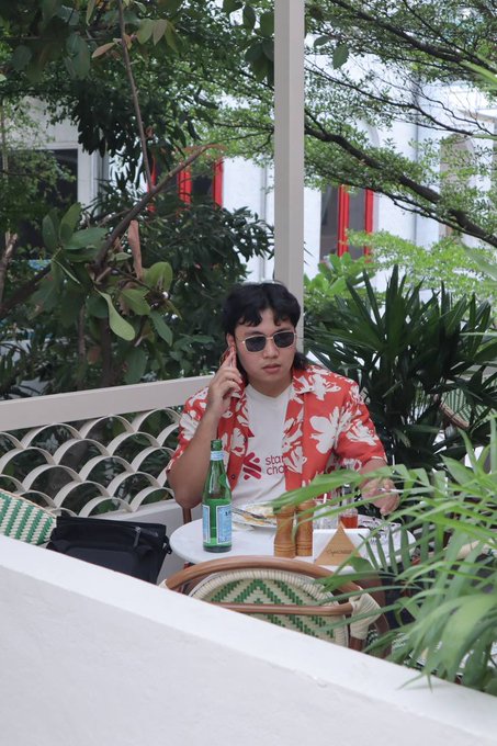 A person wearing a red Hawaiian shirt, sunglasses, and a white t-shirt sits at an outdoor cafe table with drinks and food, surrounded by lush greenery.
