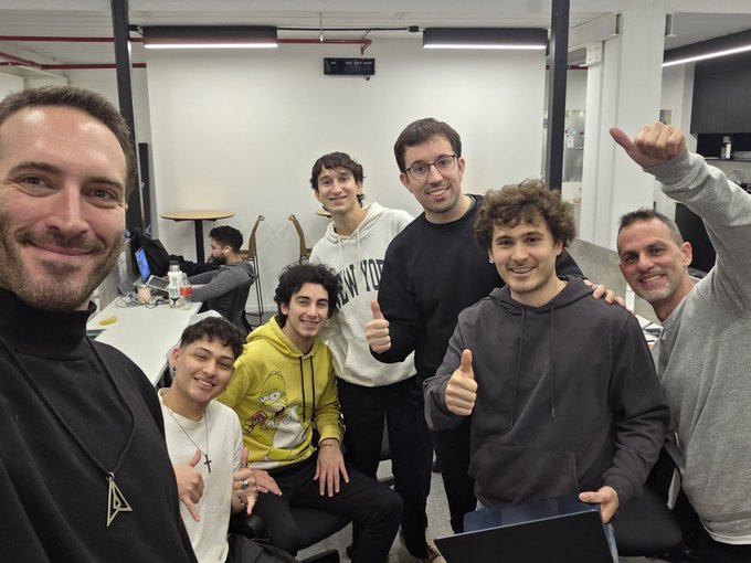 The image features a group of six young men posing for a photo with their arms raised in a friendly manner. They are all wearing hoodies and have laptops open on their laps, suggesting that they might be working together or sharing ideas. The background includes a white wall and a desk, indicating that the setting is likely an office environment. The post about AI agents being turned into real market participants seems to be related to the group of young men in the image, as it could imply that they are part of a team working on building AI agents or competing under constraints using AI technology.