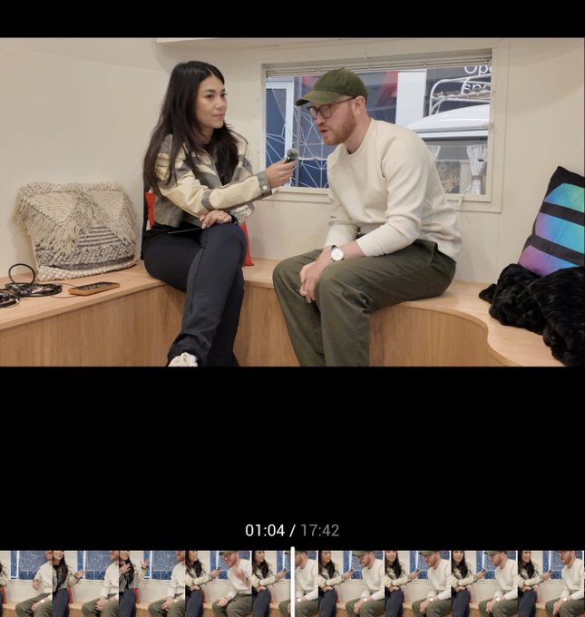 A video frame at 01:04/17:42 showing a woman interviewing a seated man wearing a green cap and glasses inside what appears to be a trailer.