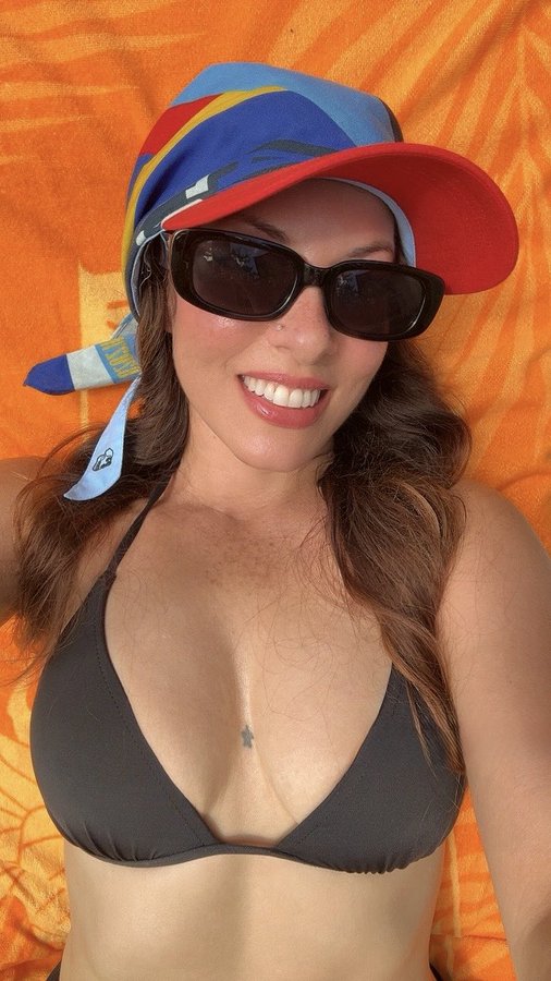 A smiling woman wearing a colorful hat, sunglasses, and a dark bikini top poses in front of an orange patterned background.