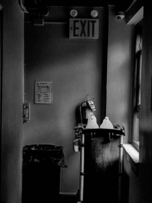 A black and white photograph of a dimly lit room featuring an illuminated 'EXIT' sign above, with fire extinguishers, a fire extinguisher inspection certificate on the wall, and a window to the right.