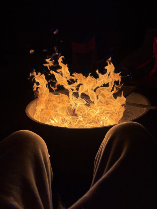 A first-person view of someone sitting by a fire pit with bright orange flames, with their knees visible in the foreground and a dark night setting around them.