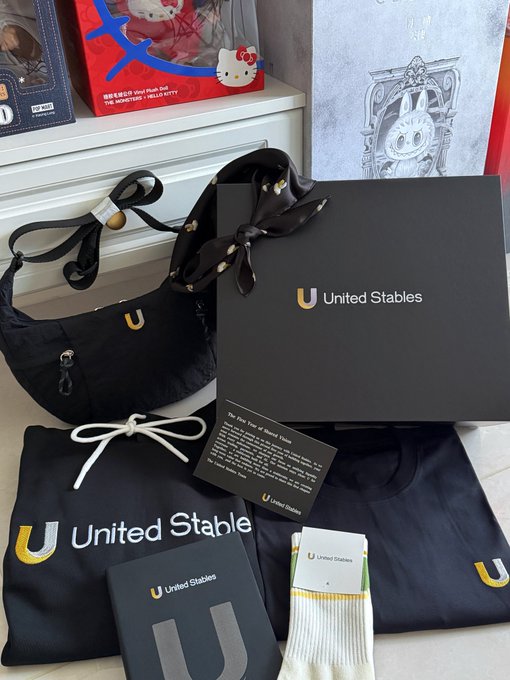 A display of "United Stables" merchandise, including a black bag with a "U" logo, a black drawstring pouch, a neckerchief, and socks, alongside other pop culture-themed boxes.