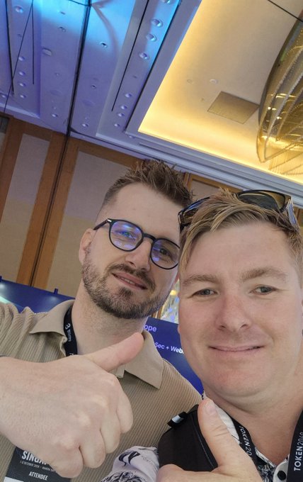 A selfie of two men at what appears to be a conference, both giving thumbs up, with one wearing glasses and a beige shirt with a 'Silicon' badge, and the other wearing sunglasses on his head with a 'TOKEN2049' lanyard.