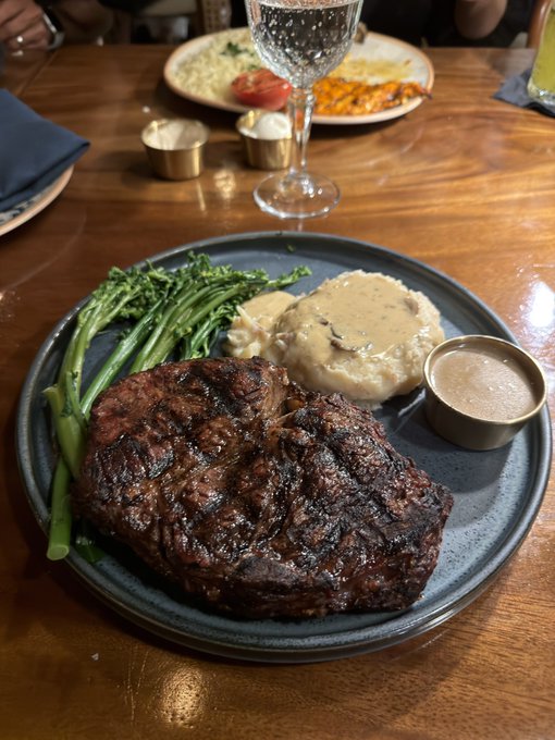A perfectly grilled ribeye steak is served with broccolini, mashed potatoes, and au jus on a dark plate, with a wine glass and side dishes visible on the wooden table. The caption "Minted this for 0.03 eth - Did i get rugged?" humorously contrasts the high-end steakhouse meal with the user's concern about a potentially failed NFT mint or crypto investment at an extremely low price.
