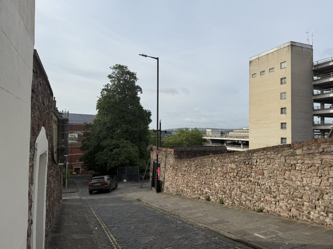 The image shows a street with a tall building, possibly an office or residential structure, on one side and a brick wall on the other. A car can be seen parked near the bottom of the photo. The sky appears cloudy, suggesting that it might be a slightly overcast day. The post in the image reads "GM Bristol", which could potentially be related to the location of the building or an event taking place nearby.