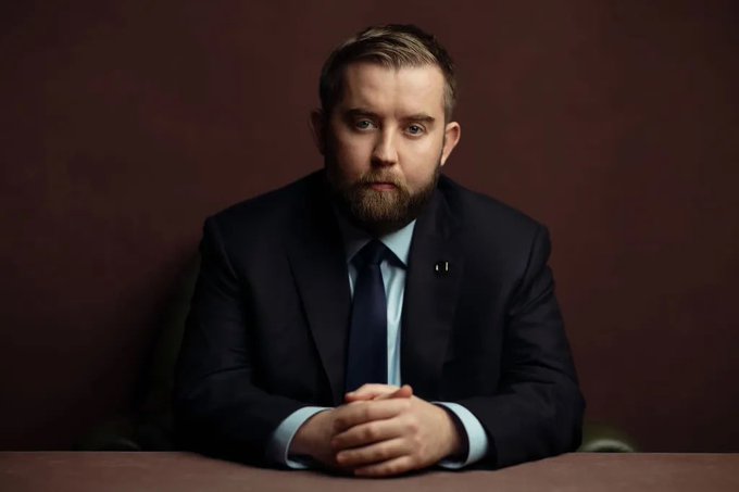 A bearded man in a dark suit and tie sits at a table with his hands clasped, gazing directly at the camera against a dark maroon backdrop. His serious, contemplative expression conveys the gravity of the enormous financial scale suggested by the post's reference to "Quadrillions."
