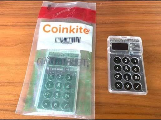 A physical Coinkite hardware wallet with a numeric keypad sits next to its clear plastic anti-tamper packaging on a wooden table.