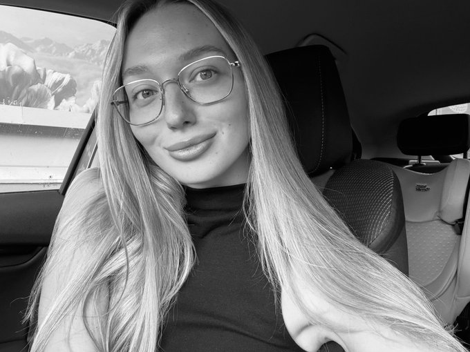 A black-and-white selfie of a young woman with long blonde hair and wire-framed glasses, smiling from inside a car with a snowy mountain landscape visible through the window.