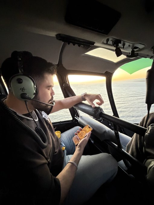 A man wearing a headset sits inside a helicopter flying over water at sunset, holding a snack.