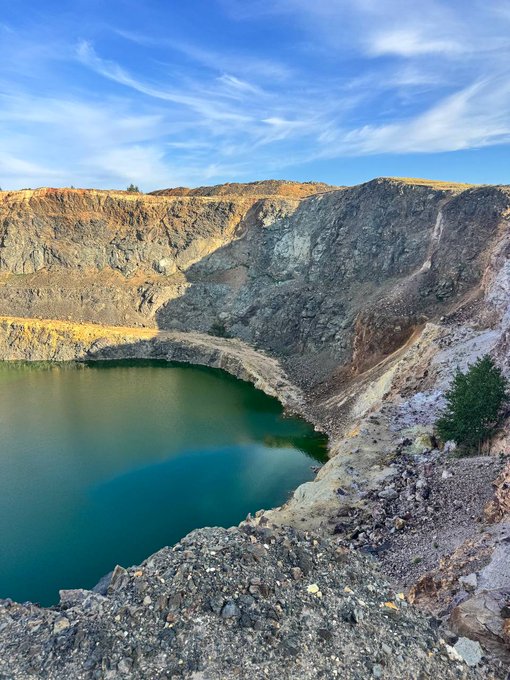 A landscape photograph of a turquoise crater lake surrounded by rocky, mineral-stained cliff walls under a blue sky with wispy clouds.