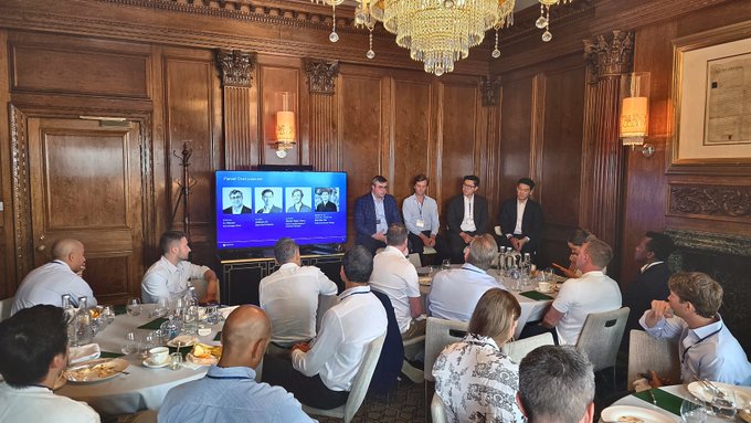A group of people in business attire seated around a dining table in an ornate wood-paneled room with a chandelier, watching a presentation on a screen titled 'Public Limited Networking' showing profiles of men in suits.