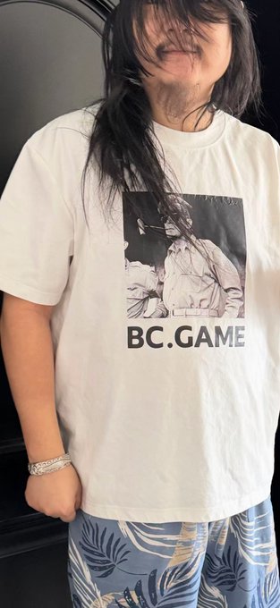 The image features a person wearing a white t-shirt with a picture of a man's face on it, along with the text "BC Game". The person appears to be posing for a photo or selfie while standing against a black background.