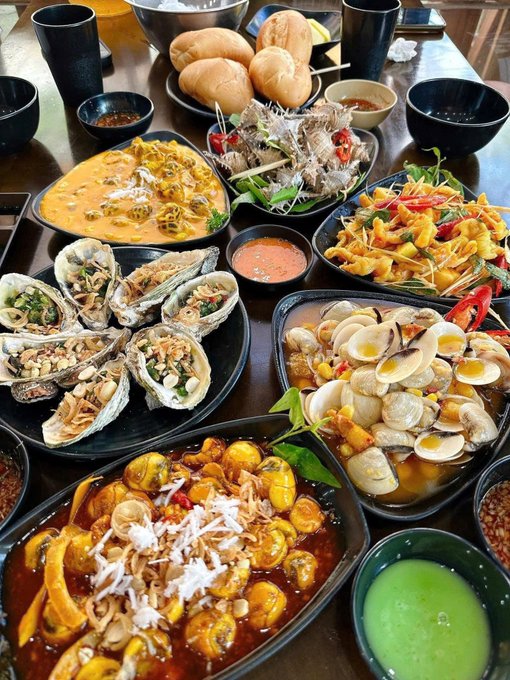 A table is covered with a lavish spread of Vietnamese dishes, including oysters topped with peanuts, clams, braised meat, and various salads and dips.