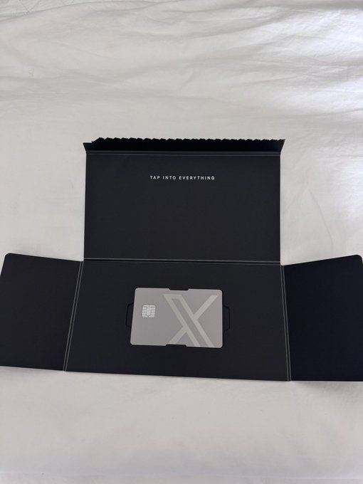 An open black presentation box on white fabric displays a silver card with 'X' on it, with the text 'TAP INTO EVERYTHING' printed on the inside lid.