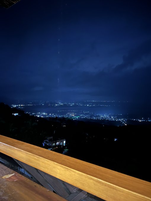 Nighttime view from a wooden balcony overlooking a city with scattered lights under a dark blue cloudy sky.