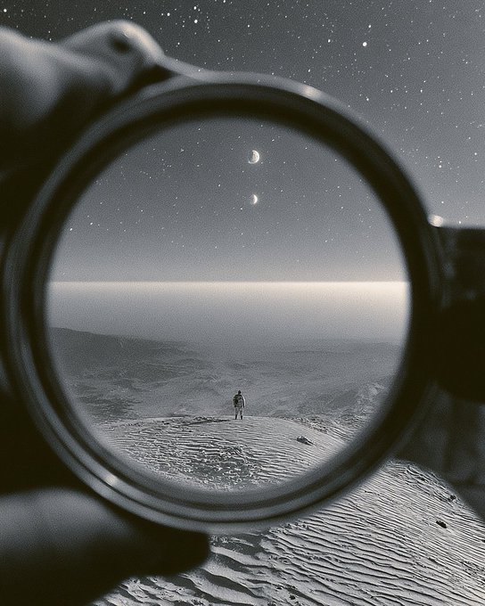 A black-and-white photo shows a hand holding a camera lens, through which a lone figure is seen standing on a sandy dune beneath a starry sky with two visible planets or moons.