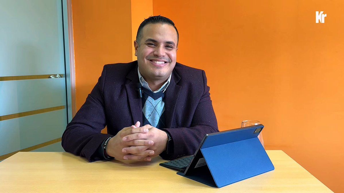 The image features a man sitting at a desk with his arms crossed while smiling for the camera. The background of the photo shows an orange wall, which contrasts nicely with the man's blue shirt. There are also two cups placed near him on the table. The overall atmosphere suggests that Jose is in a relaxed and comfortable environment as he enjoys his time at work or perhaps taking a break from it.