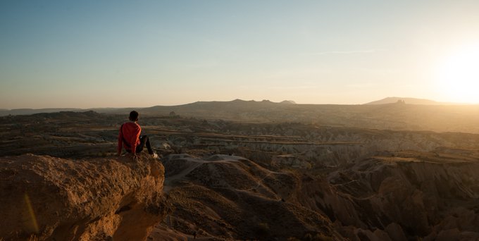 The image shows a person sitting on a rock, looking out over a desert landscape at sunrise or sunset. The person appears to be wearing a red shirt and has their back turned towards the camera. The scene captures the beauty of nature as the sun sets behind the mountains in the background.