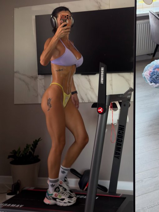 A mirror selfie of a muscular woman in a lavender bikini top and yellow bottoms taking a photo with her phone while wearing headphones in a gym setting.
