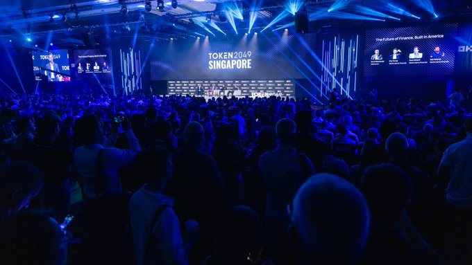 A large conference venue illuminated in blue light with screens displaying 'TOKEN 2049 SINGAPORE' to a massive seated audience.