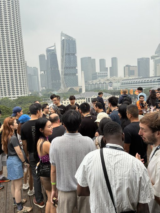 An outdoor networking event with a crowd of casually dressed attendees gathered on a wooden deck overlooking the Singapore skyline, featuring distinctive buildings including the twisting Sangenjaya-inspired tower in the city center.