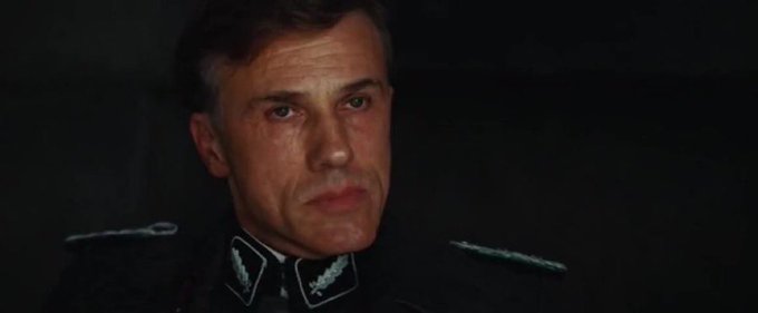 A close-up shot of a stern-looking man in a black military-style uniform with decorative collar insignia, his expression intense as he gazes slightly off-camera against a dark background. The image appears to be a still from a film, likely used to represent the serious and dramatic nature of the DOJ's insider trading charges against Robinhood employees on Hyperliquid.