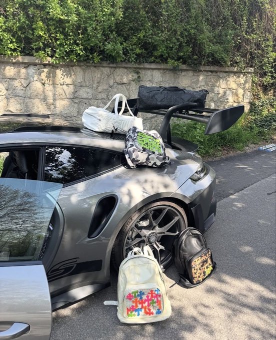 A grey Porsche 911 GT3 RS is parked on the side of a road with its door open, featuring a large rear wing and surrounded by several bags, including a white duffel bag, a camouflage backpack, and others resting on the car and the ground.