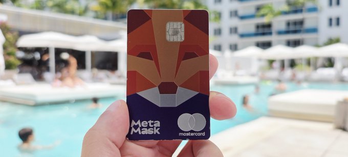 A hand holds a MetaMask Mastercard credit card with a geometric lion design, with a swimming pool and people relaxing in the background.
