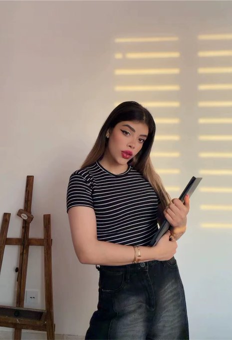 A young woman poses confidently in a striped black and white top and dark jeans, holding what appears to be an artist's portfolio or sketchbook, embodying the creative spirit suggested by the caption "Just an artist being an artist."