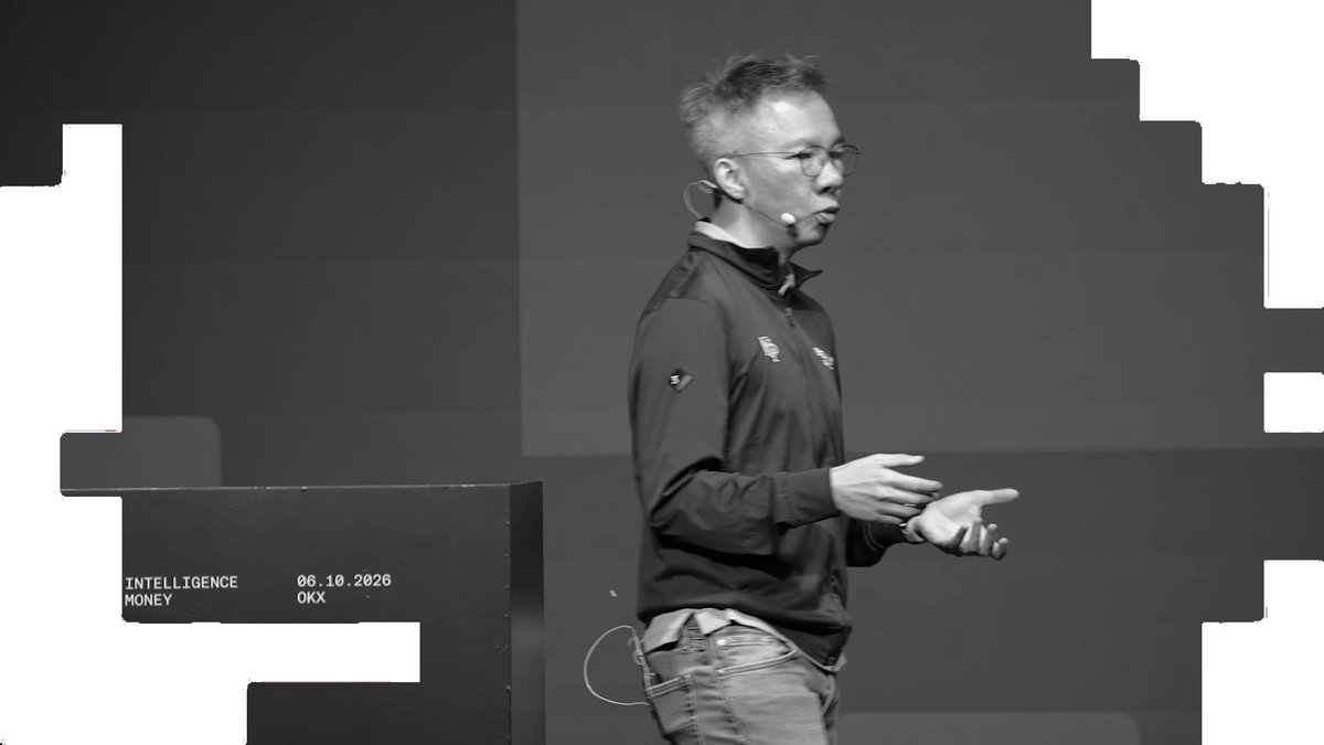 A man wearing a headset microphone and a "Ripple" branded jacket speaks on stage at an OKX event, gesturing as he addresses the audience. The backdrop displays text including "INTELLIGENCE," "MONEY," the date "06.10.2026," and "OKX," aligning with the post's announcement of OKX expanding its offerings across multiple markets.