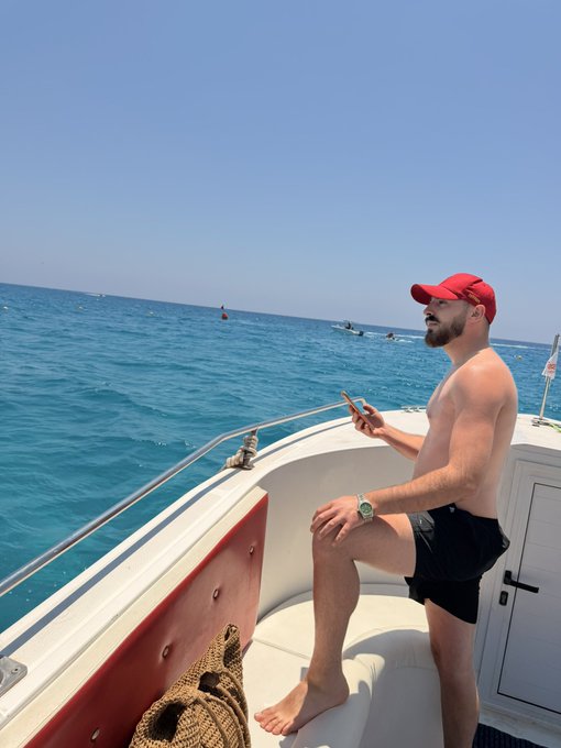 A shirtless man in a red cap and black shorts sits on a boat's bow, scrolling on his phone while looking out at the turquoise sea on a bright sunny day. The caption "Gm friends" pairs well with the relaxed morning vibes of a leisurely boat ride.