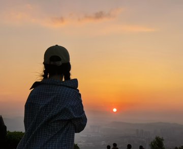 A person in a checkered jacket and cap watches the sunrise over a hazy cityscape, capturing the reward of waking up at 4 AM to witness the dawn break.
